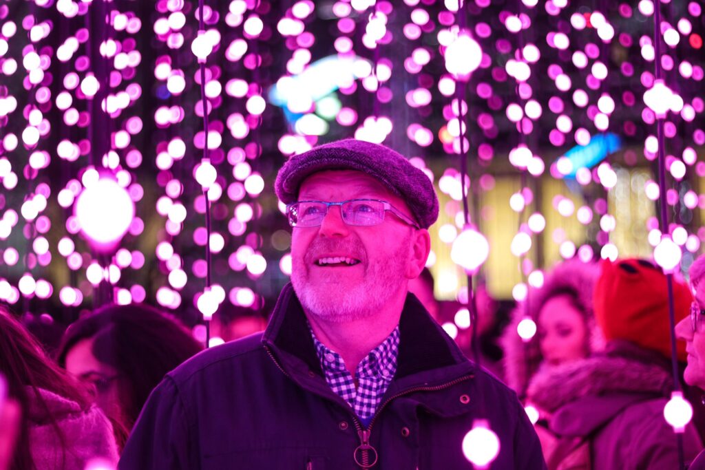 Un homme portant des lunettes, une casquette plate et une veste sourit, debout parmi des guirlandes lumineuses roses, entouré de gens dans un environnement vibrant et lumineux  - hote airbnb senior