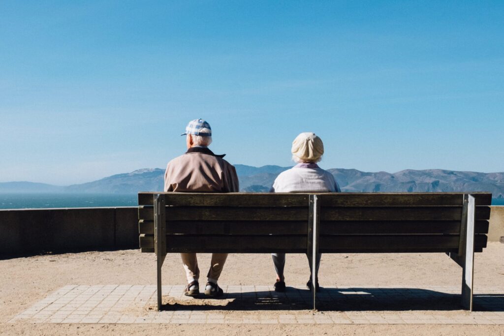 Deux personnes âgées sont assises côte à côte sur un banc, face à une vue panoramique sur les montagnes et l'eau sous un ciel bleu clair - hote airbnb senior