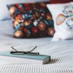 A closed book with eyeglasses resting on top lies on a striped bedspread, with two colorful, patterned pillows in the background - student airbnb rentals