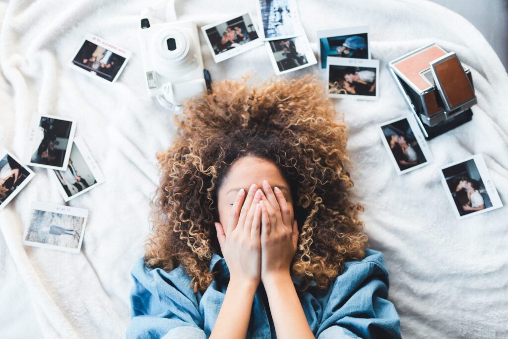 Une personne aux cheveux bouclés est allongée sur un lit et se couvre le visage avec les mains, entourée de photos instantanées éparpillées - annonce airbnb instagram