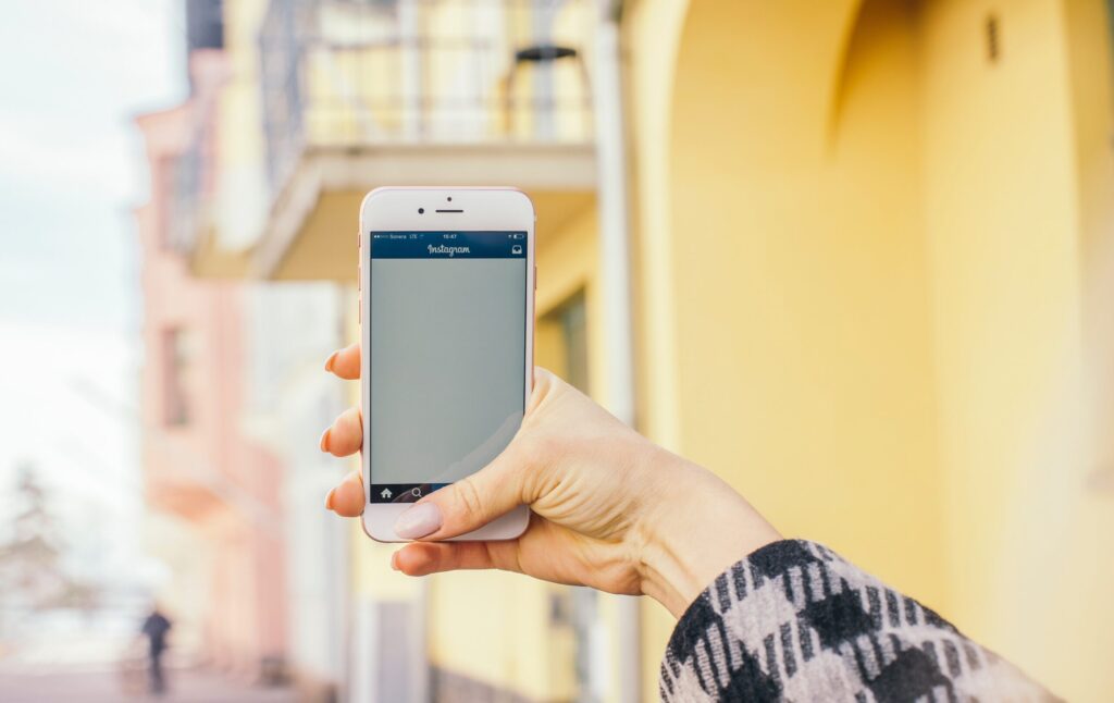 Une personne brandit un iPhone affichant l'application Instagram, alors qu'elle se trouve à l'extérieur, près de bâtiments aux couleurs pastel et aux murs jaunes - annonce airbnb instagram