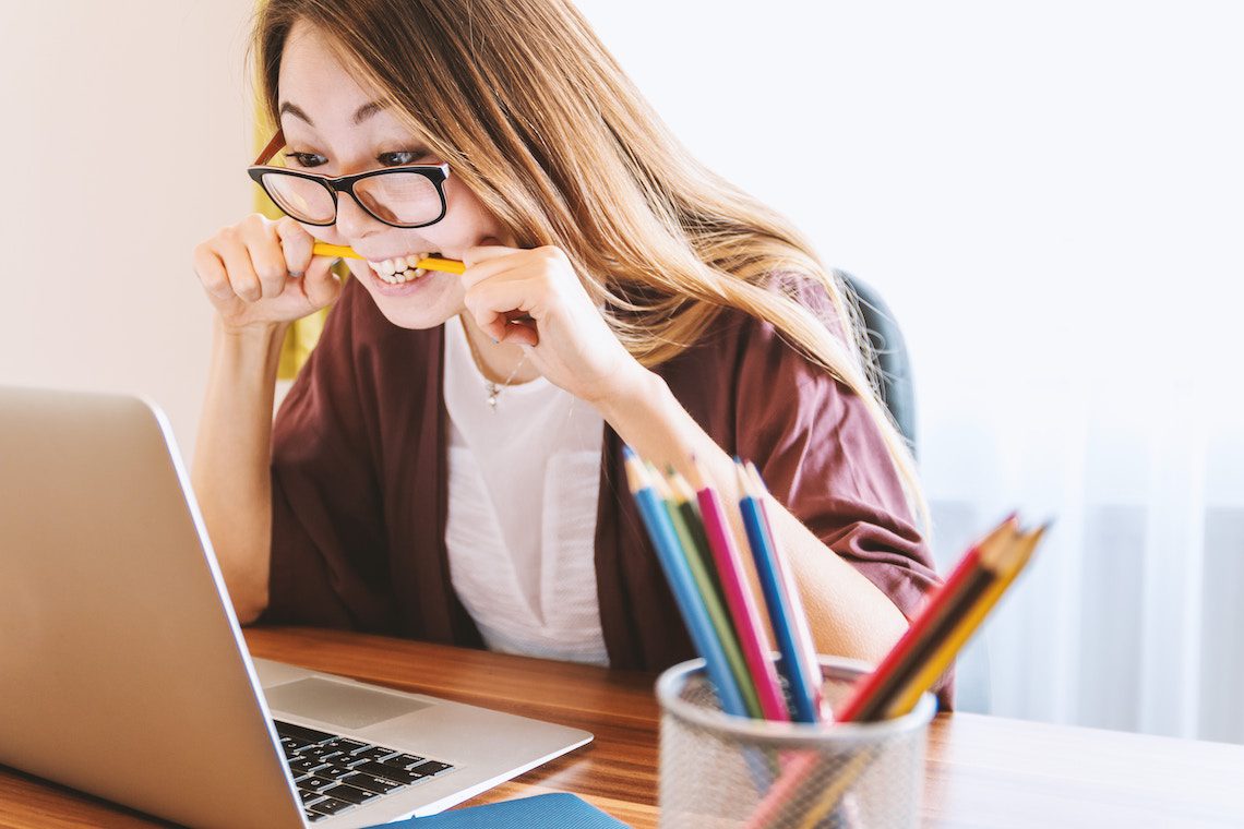 Une jeune femme portant des lunettes est assise à un bureau, mordant un crayon avec frustration tout en regardant un ordinateur portable. Une boîte de crayons de couleur et un carnet de notes sont posés sur le bureau. L'arrière-plan est clair et légèrement éclairé - airbnb résidence principale