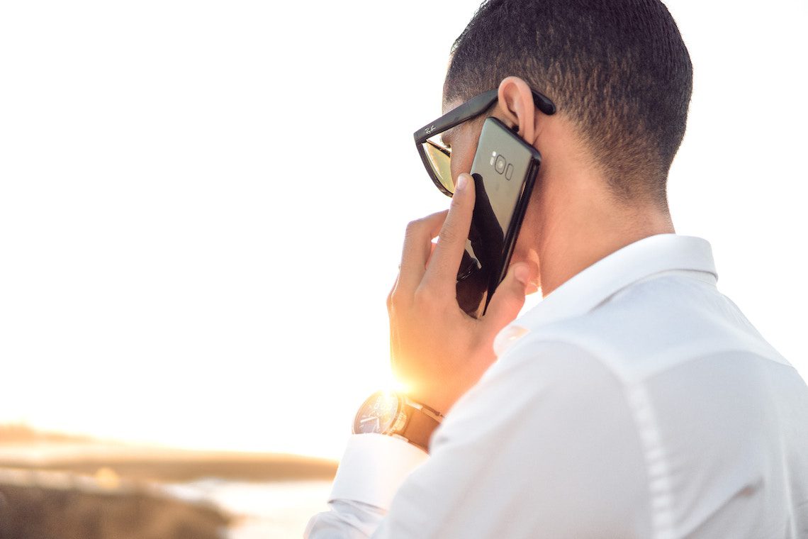 Un homme portant des lunettes de soleil et une chemise blanche se tient à l'extérieur, tenant un smartphone à l'oreille et parlant au téléphone - airbnb résidence principale