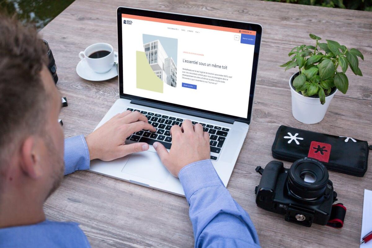 Une personne tape sur un ordinateur portable à une table en bois avec une tasse de café, un appareil photo, un étui et une plante en pot à proximité - meilleurs logiciels de location saisonnière