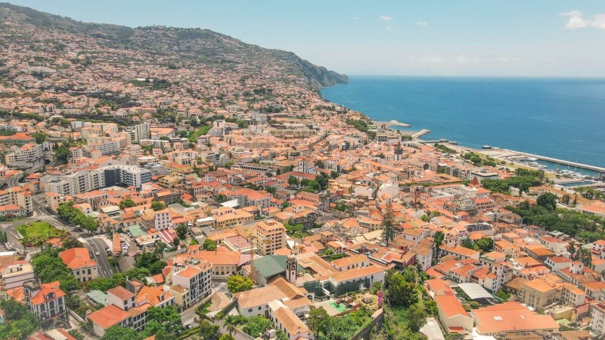 Aerial view of a coastal city with many red-roofed buildings, lush greenery, and hills in the background - buying property in madeira