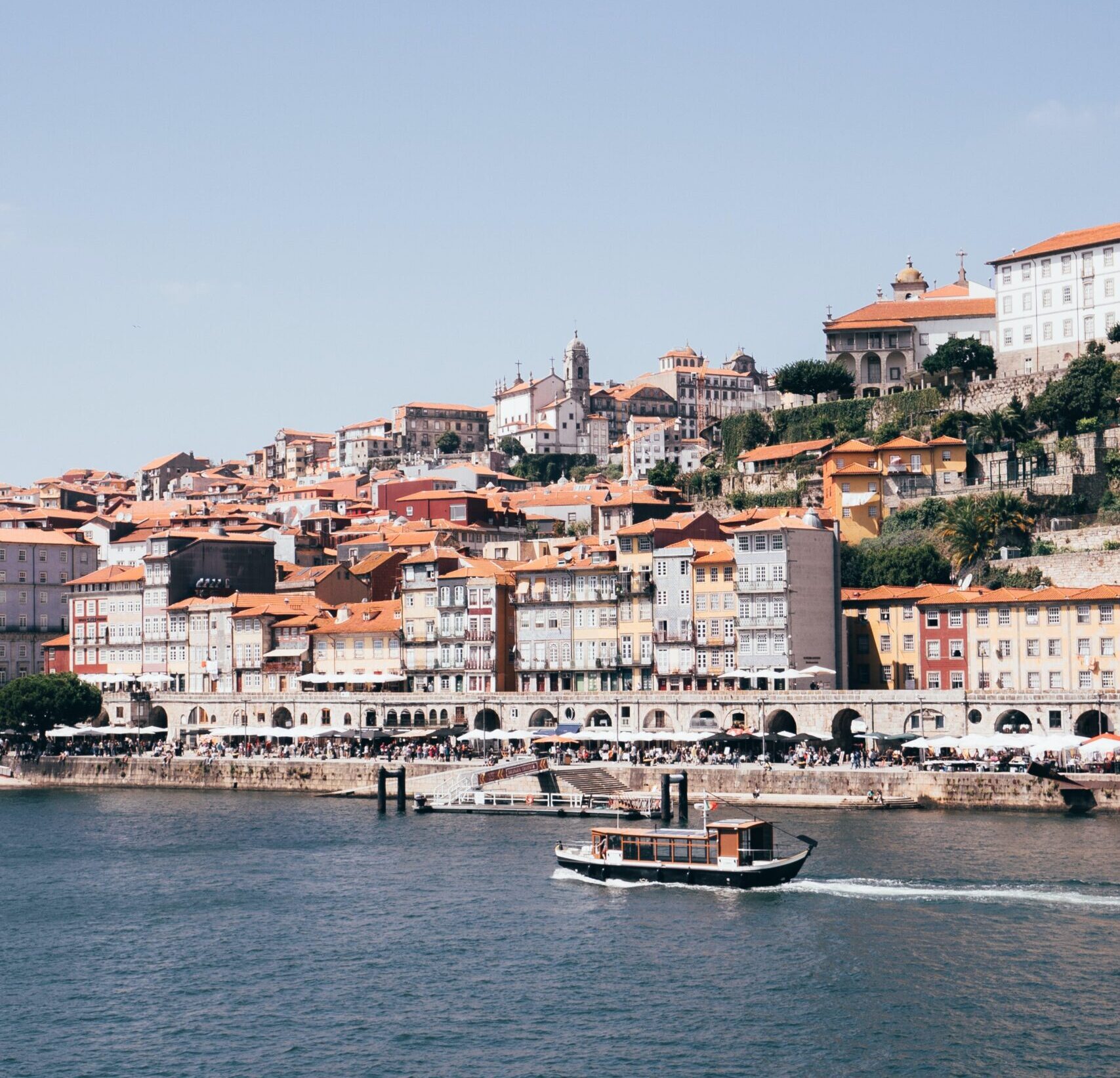A small boat moves along the Douro River with Porto’s colorful hillside buildings in the background - Porto city break