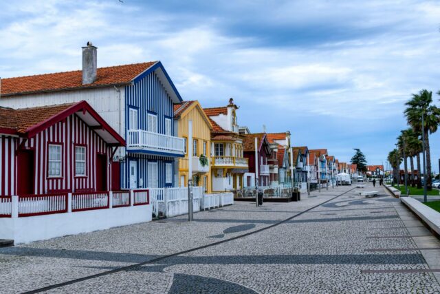 A row of colorful striped beach houses with red, blue, yellow, and white facades stands along a wide, patterned stone promenade. The houses have red-tiled roofs, balconies, and white shutters. Palm trees line the right side of the walkway under a cloudy blue sky - where to go on a holiday in january