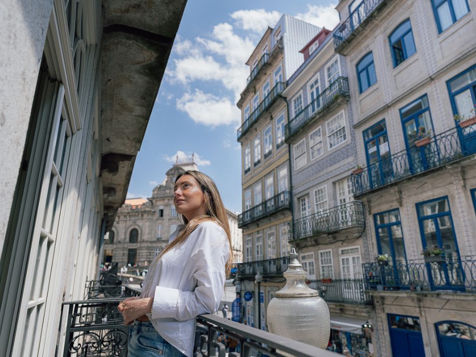 A woman stands on a balcony at the GuestReady apartment in Oporto Cardosas, looking up at the blue sky, with historic tiled buildings and iron balconies lining the street below.