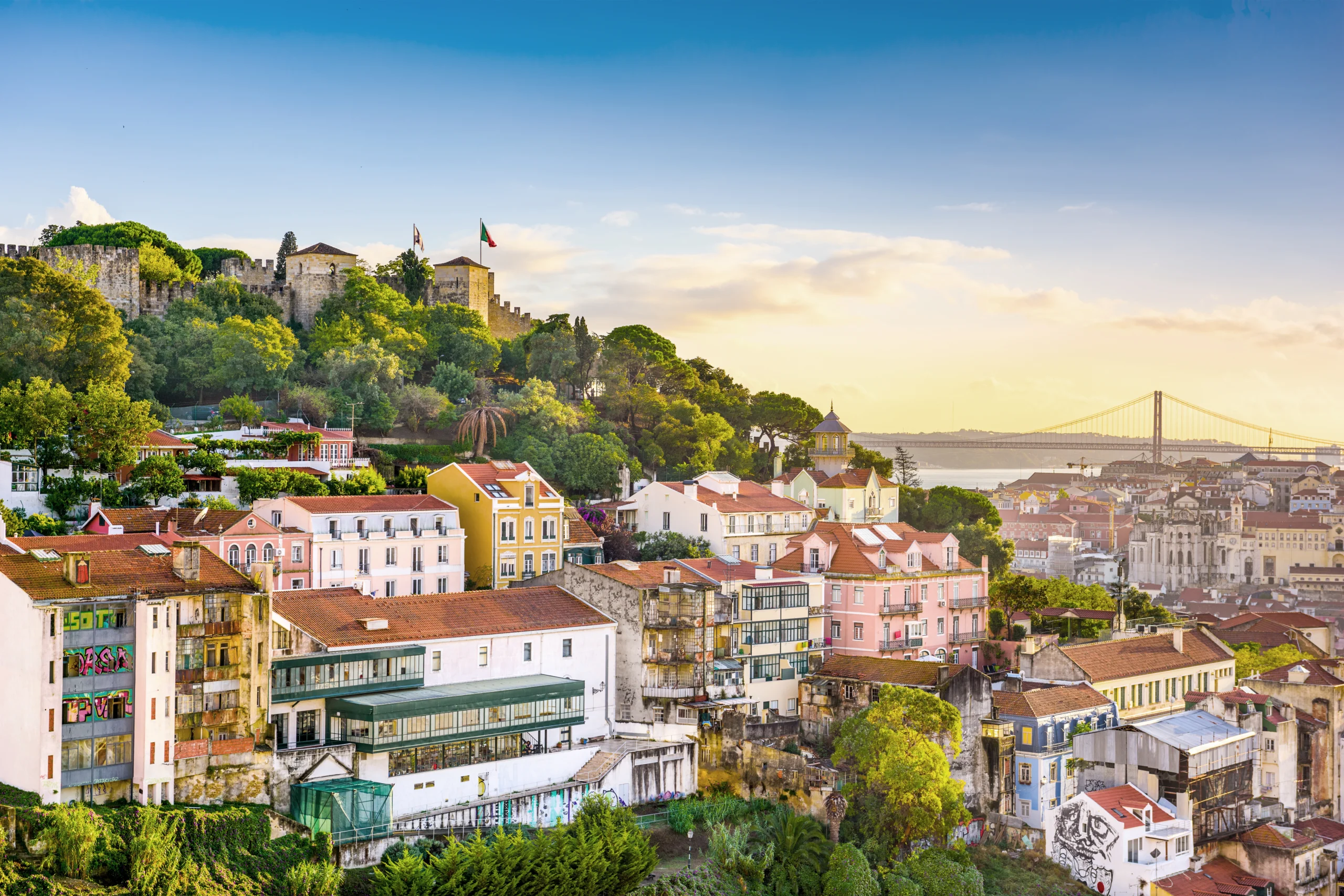 Colorful hillside houses in Lisbon with São Jorge Castle and the 25 de Abril Bridge in the background - invest in Lisbon