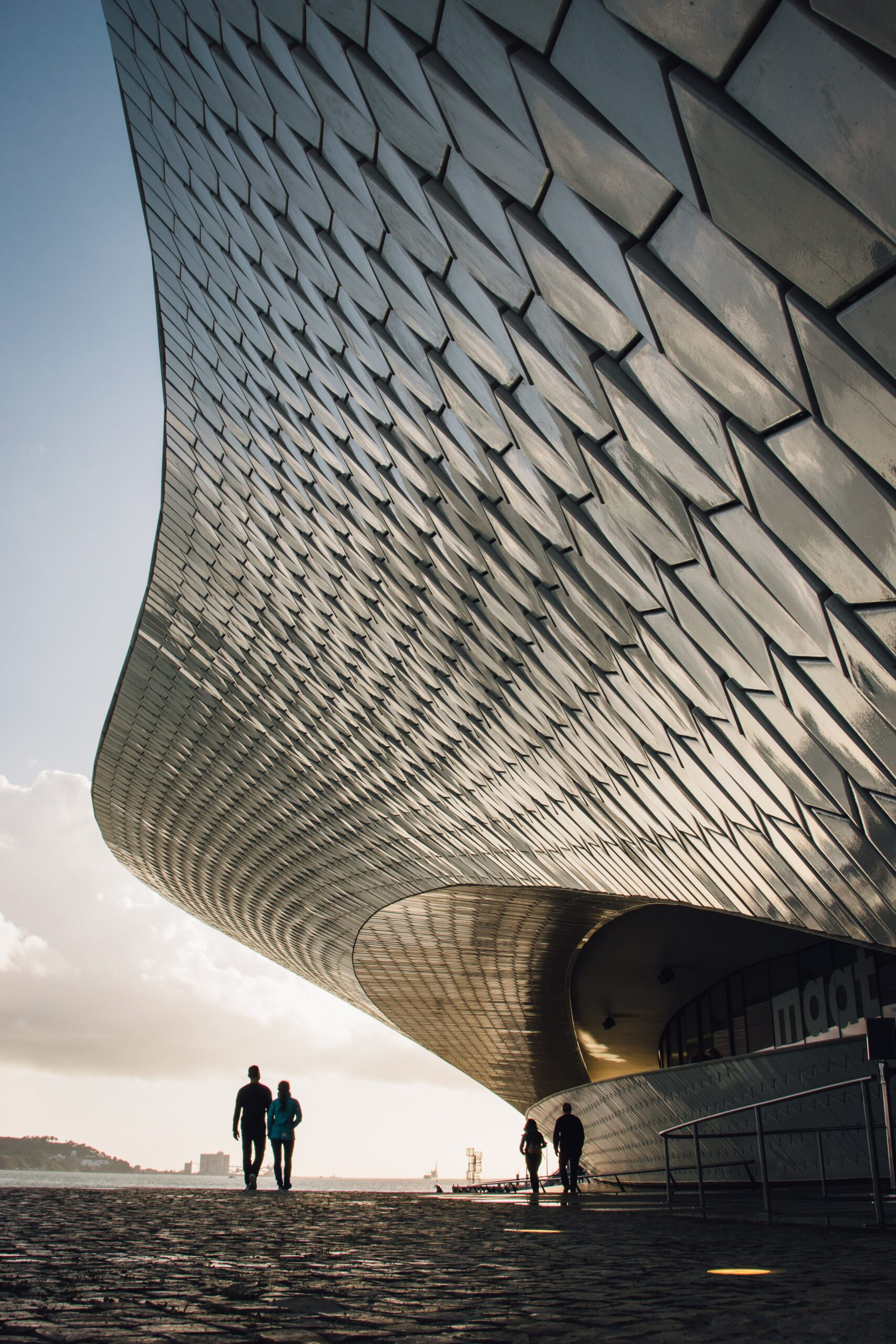 People walking beside the curved modern architecture of the MAAT museum along Lisbon’s waterfront - invest in lisbon.