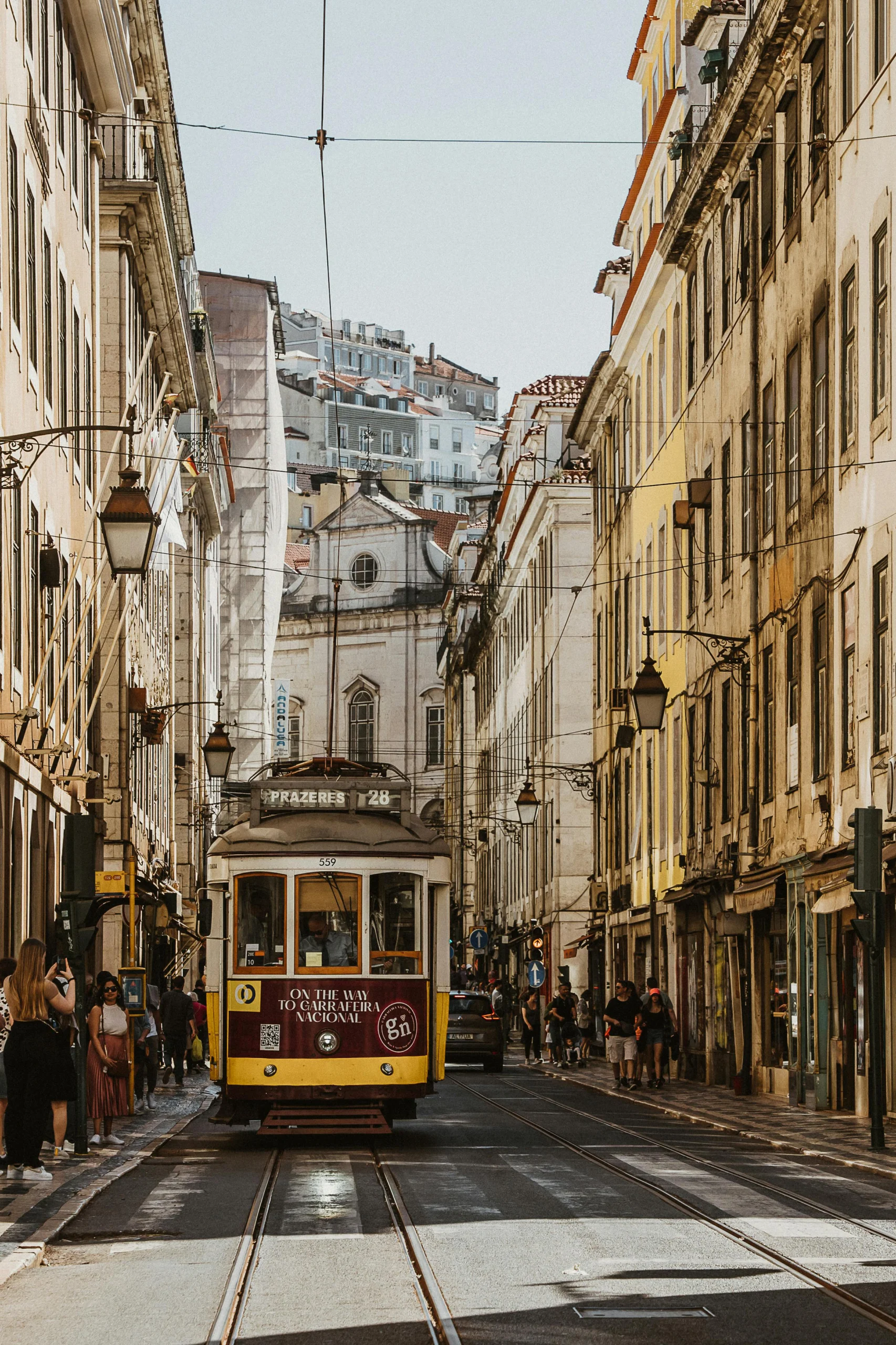 Classic yellow Lisbon tram traveling through a narrow historic street lined with buildings and pedestrians - invest in Lisbon