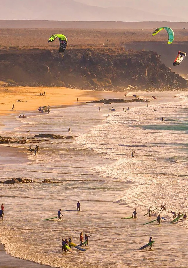 Kitesurfers and beachgoers along a golden sandy beach on Fuerteventura’s rugged coastline - GuestReady Airbnb management in Fuerteventura