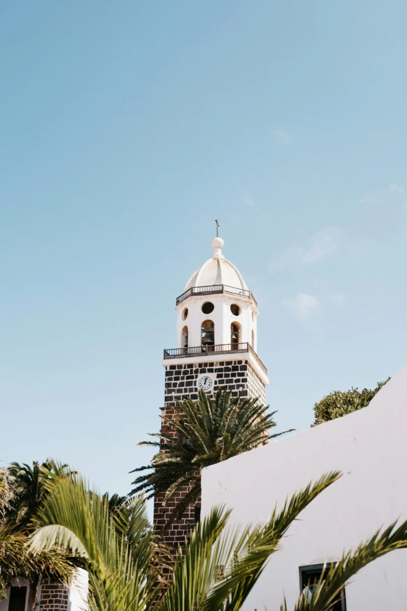 White church bell tower with a clock, framed by palm trees under a clear blue sky in Lanzarote