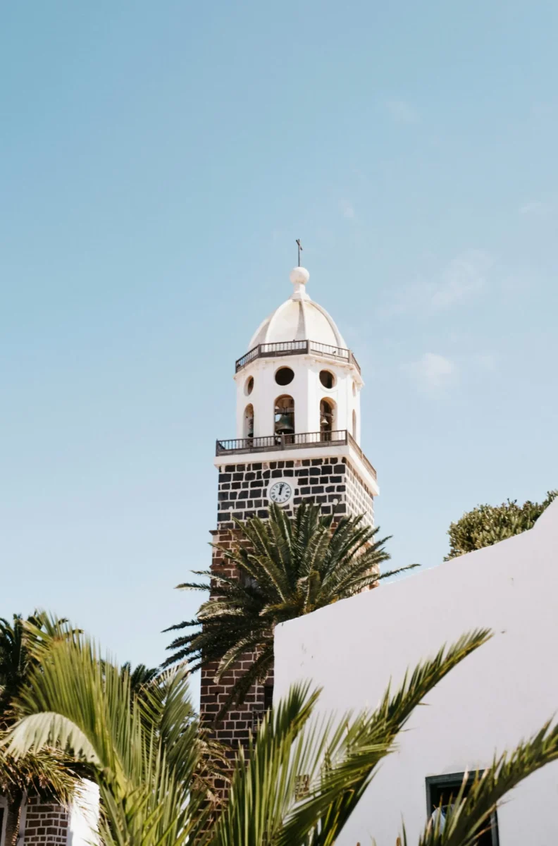 White church bell tower with a clock, framed by palm trees under a clear blue sky in Lanzarote