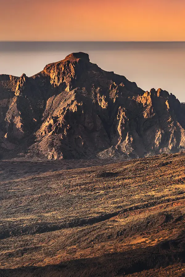 Montaña volcánica rocosa al atardecer, con luz cálida y un horizonte brumoso al fondo.