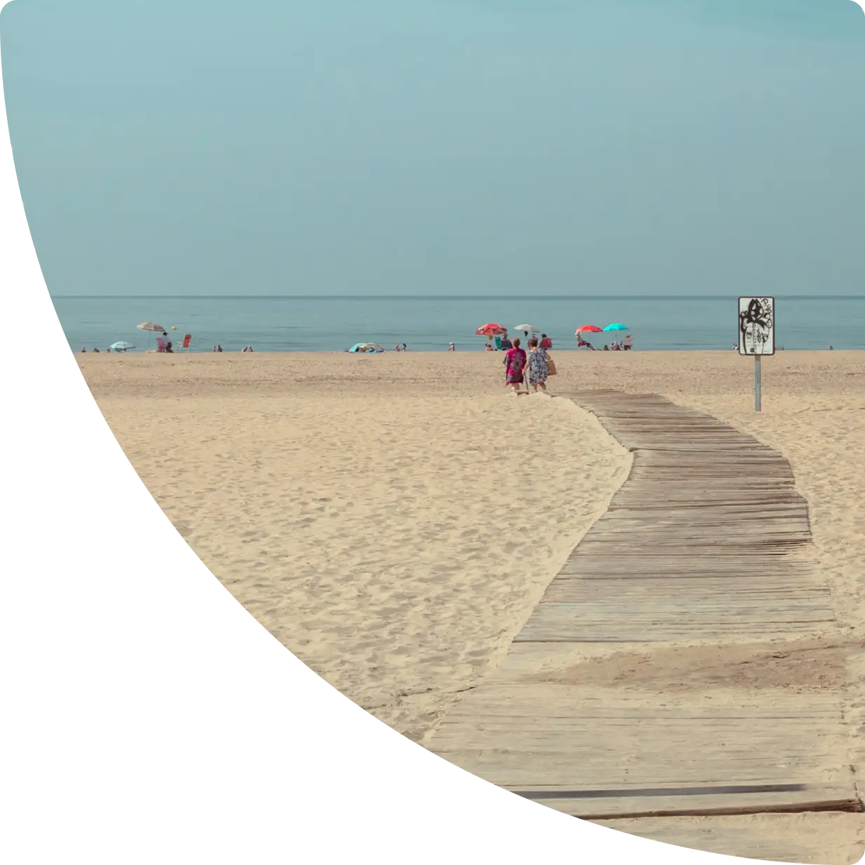 Wooden boardwalk across sandy dunes to a calm beach with umbrellas and people by the shore - Airbnb management in Huelva, GuestReady