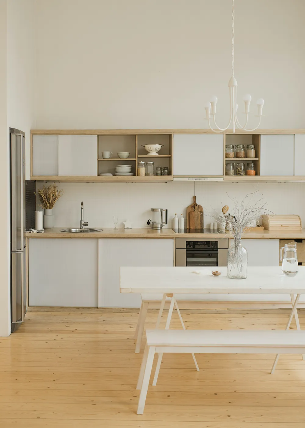 Cocina minimalista con muebles de madera clara, estanterías abiertas y mesa de comedor blanca.